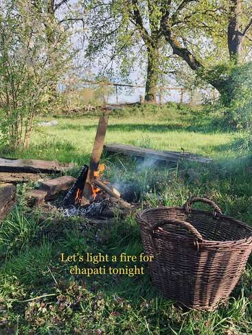 A cozy outdoor scene with a small campfire, green grass, blooming trees, and an empty wicker basket. Text reads: “Let’s light a fire for chapati tonight.”