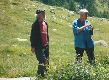 L'image représente deux personnes debout dans un champ herbeux, entouré de rochers et d'arbres, l'une d'elles tenant un petit objet à la main.