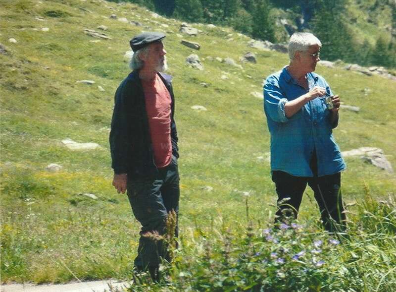 The image depicts two individuals standing in a grassy field, surrounded by rocks and trees, with one person holding a small object in their hand.