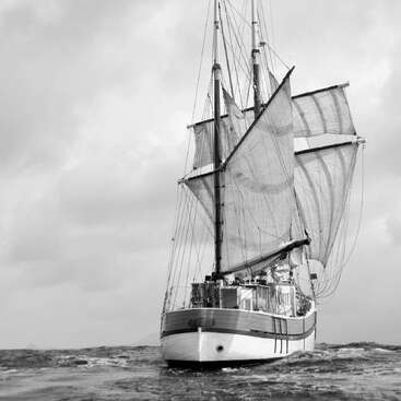 The image depicts a sailboat with three sails on a body of water, set against an overcast sky, showcasing its majestic presence.