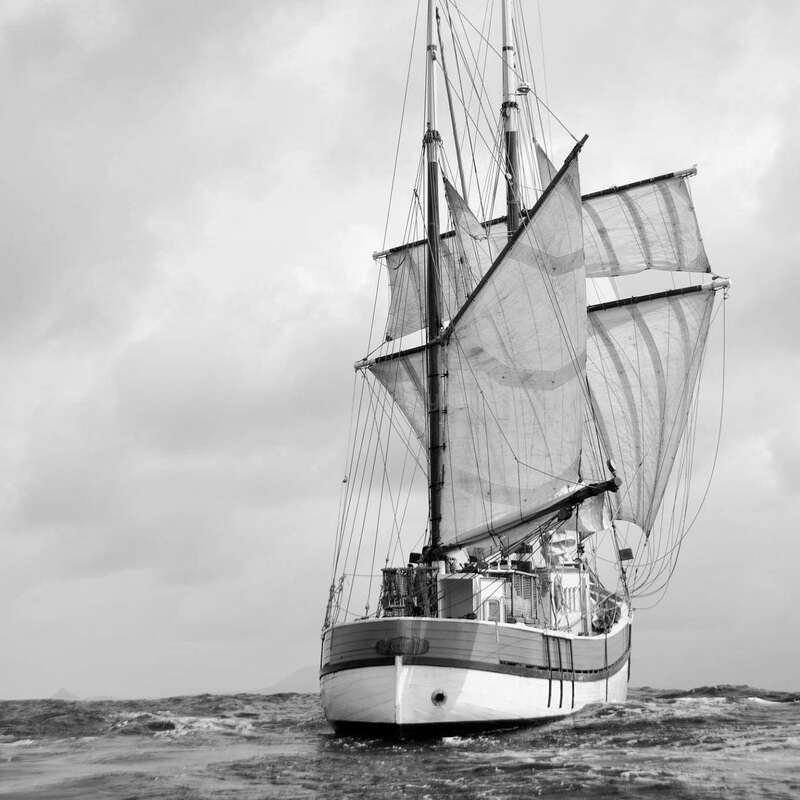 L'image représente un voilier à trois voiles sur un plan d'eau, dans un ciel couvert, mettant en valeur sa présence majestueuse.