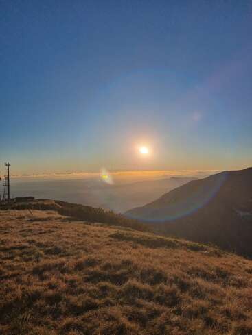 Ein wunderschöner Sonnenaufgang oder Sonnenuntergang über einer Berglandschaft, bei dem die Sonne über den Hügeln glüht, einen Linsenreflex erzeugt und warmes Licht auf grasbewachsenes Terrain wirft.