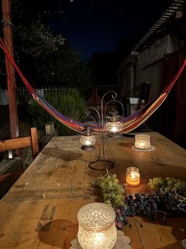 Ein rustikaler Holztisch steht nachts im Freien, geschmückt mit brennenden Kerzen, Weintrauben und Laternen. Im Hintergrund spannt sich eine bunte Hängematte.