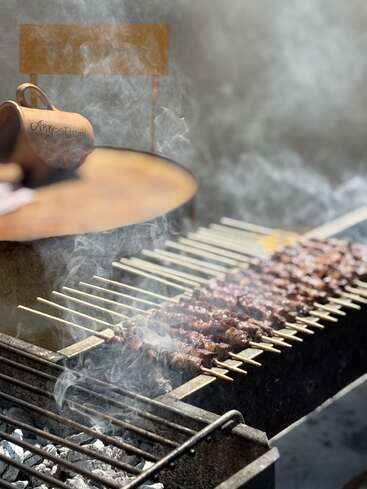 Fleischspieße werden über heißen Kohlen gegrillt, Rauch wirbelt durch die Luft. Im Hintergrund sorgen ein rustikaler Tisch und ein Keramikbecher für Charme.