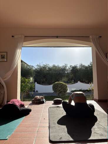 Zwei Menschen machen drinnen Yoga unter einem offenen Torbogen, durch den Sonnenlicht hereinströmt. Eine Katze ruht sich in der Nähe auf Decken aus, umgeben von Grün und einer friedlichen Landschaft.