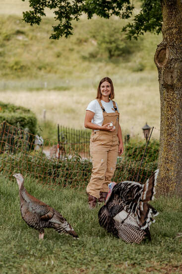 Una mujer con un mono marrón claro y una camisa blanca está de pie al aire libre, sonriendo mientras sostiene unos huevos. Dos pavos deambulan cerca, con árboles y una valla detrás de ella.