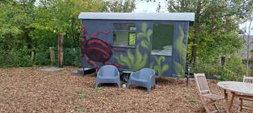 Una pequeña y artística caravana con un mural de temática natural está rodeada de árboles. Delante, dos sillones grises y una mesa de madera con sillas invitan a relajarse.
