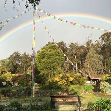 La imagen representa una serena escena de jardín con un vibrante arco iris, un banco de madera y un mástil adornado con banderas de colores, sobre un fondo de frondosos árboles y cielo gris.