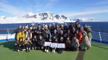 Eine große Gruppe posiert zusammen auf einem Schiffsdeck mit verschneiten Bergen und blauem Meer im Hintergrund, alle in Winterkleidung, lächelnd und fröhlich.