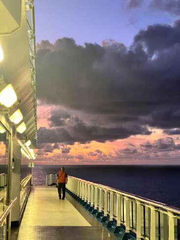 Eine Person geht bei Sonnenuntergang allein auf einem hell erleuchteten Schiffsdeck. Dunkle Wolken ziehen auf, während sich der bunte Himmel wunderschön auf dem ruhigen Meer spiegelt.