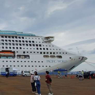 Ein großes weißes Kreuzfahrtschiff namens "Pacific World" liegt im Hafen. Mehrere Menschen stehen in der Nähe und schauen und zeigen auf das beeindruckende Schiff unter dem bewölkten Himmel.