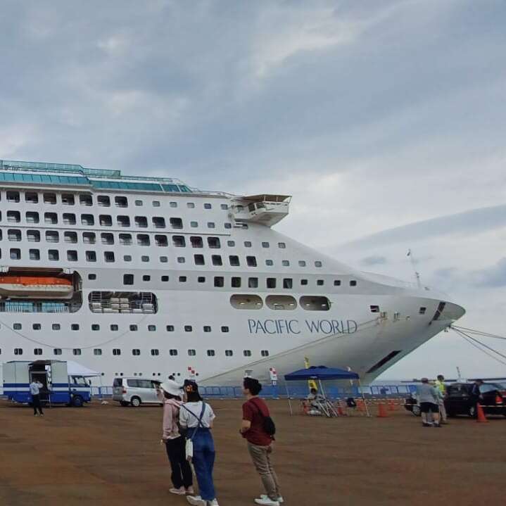 A large white cruise ship named "Pacific World" is docked at port. Several people stand nearby, looking and pointing at the impressive vessel under cloudy skies.