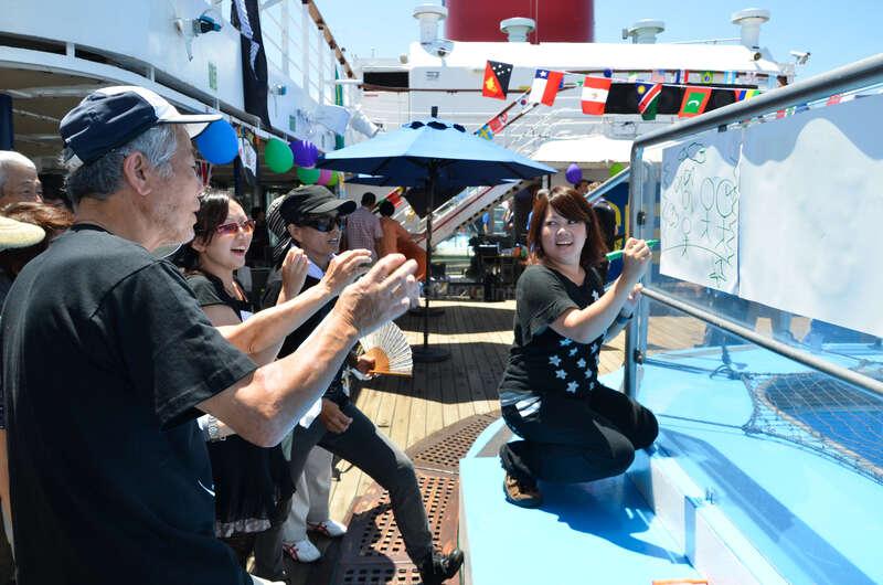 L'image représente un groupe de personnes sur le pont d'un bateau de croisière, avec une femme qui dessine sur un tableau blanc et d'autres qui applaudissent et regardent.