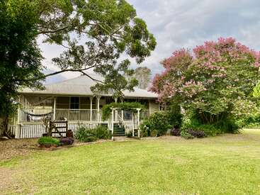 Une charmante maison de campagne avec un porche enveloppant se trouve au milieu d'une verdure luxuriante et d'arbres en fleurs. Le jardin bien entretenu et le hamac créent une atmosphère paisible.