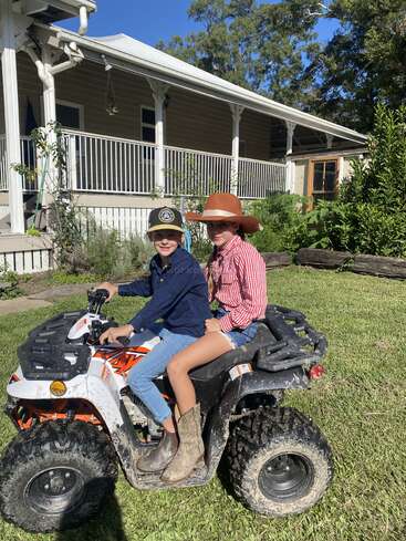 Deux enfants portant des chapeaux de cow-boy sont assis sur un VTT boueux devant une maison, profitant d'une journée ensoleillée en plein air. Tous deux sourient et portent des bottes.
