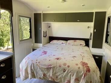 Cette image montre une chambre de camping-car confortable avec un grand lit, un couvre-lit à fleurs, des placards de pavillon, des fenêtres latérales et une lumière naturelle brillante provenant de l'extérieur.