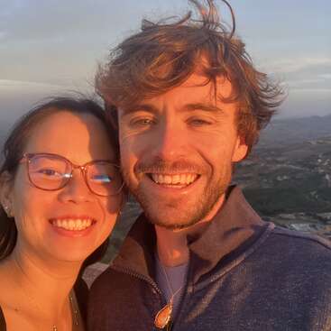 L'image montre un selfie d'un homme et d'une femme se tenant ensemble en plein air, la femme à gauche et l'homme à droite, tous deux souriant à l'appareil photo.