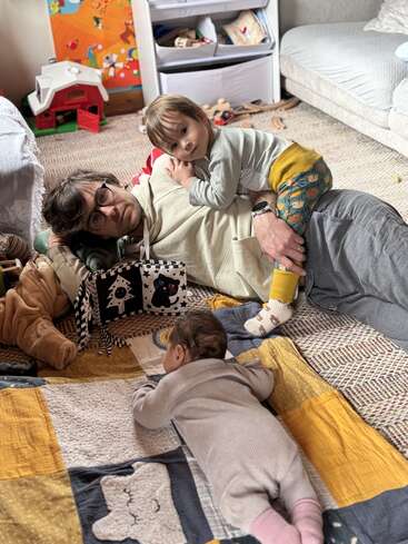 Un homme est allongé par terre, un jeune enfant sur le dos, tandis qu'un bébé est à quatre pattes à côté. Des jouets et des livres les entourent. L'ambiance est chaleureuse et ludique.