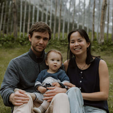 Eine junge Familie sitzt draußen auf dem Rasen und posiert für ein Foto. Der Mann und die Frau stehen zu beiden Seiten eines kleinen Kindes, und alle lächeln sanft in einer natürlichen, waldreichen Umgebung.