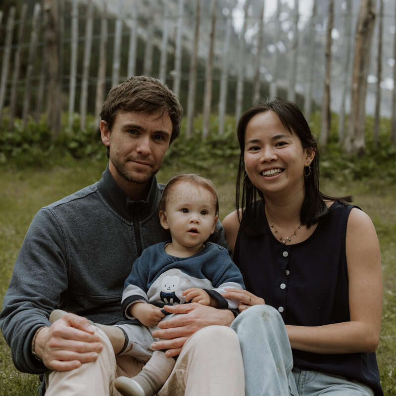 Une jeune famille est assise sur l'herbe, à l'extérieur, et prend la pose pour une photo. L'homme et la femme encadrent un petit enfant ; tous sourient doucement dans un cadre naturel et boisé.