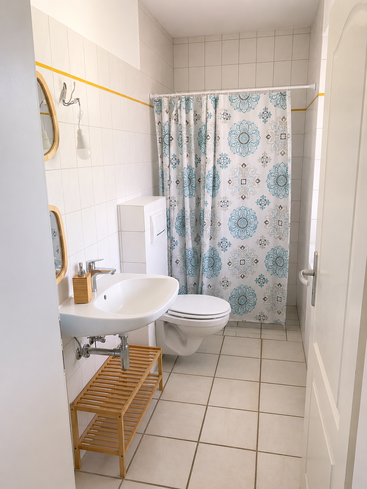 Il s'agit d'une salle de bains lumineuse et bien rangée, avec des murs et un sol carrelés, des équipements blancs, un rideau de douche à motifs, des touches de bois, des miroirs et une étagère en bois à lattes.