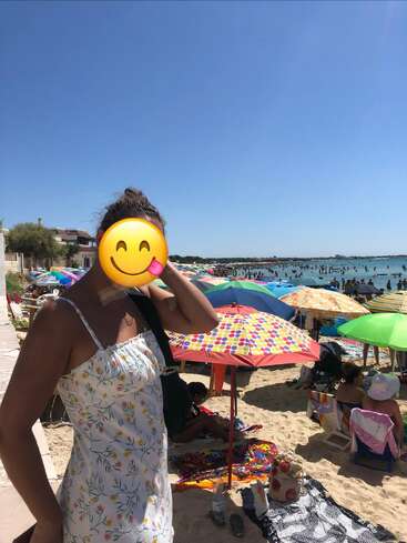 A woman in a floral dress stands on a crowded, sunny beach with colorful umbrellas. Her face is covered by a smiley face emoji. Blue sky above.