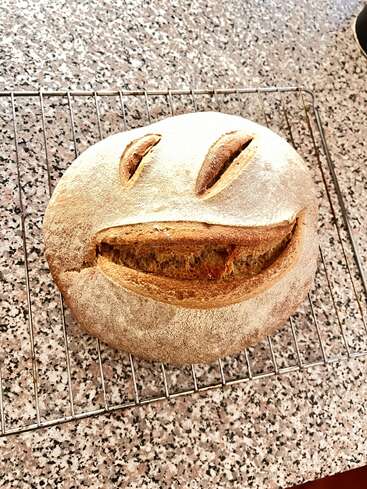 A loaf of homemade bread, cooling on a wire rack. The bread has cuts resembling a smiling face, creating a cheerful and playful look on the crust.
