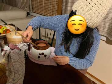A person wearing a knitted hat and blue sweater stirs a brown pot on a table, face covered by an emoji. Cozy kitchen background with fruit.