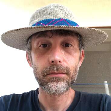 A man with a beard and mustache is wearing a straw hat with a colorful band. He has a neutral expression and is looking straight ahead.