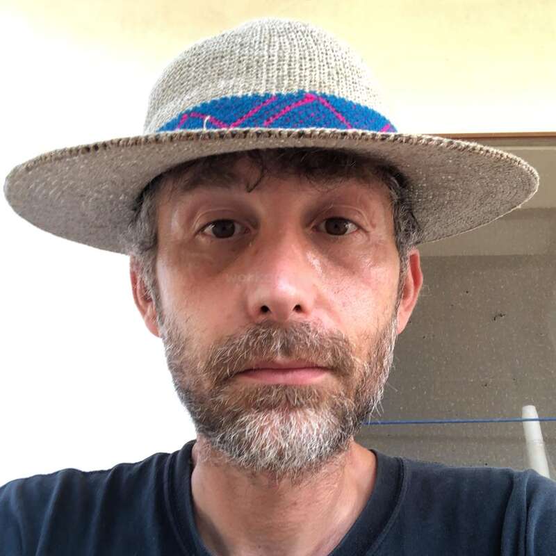 A man with a beard and mustache is wearing a straw hat with a colorful band. He has a neutral expression and is looking straight ahead.