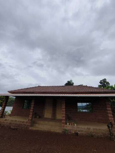 A simple brick house with a tiled roof sits under a cloudy sky. There are two doors, windows, and small potted plants on the porch.