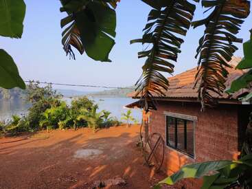 A rustic house with a tiled roof sits by a serene lake, surrounded by lush green plants and trees, with clear blue sky overhead.
