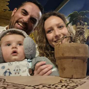 A happy family of three poses for a cheerful selfie indoors, with a baby in earmuffs, smiling parents, and a small potted plant in the foreground.
