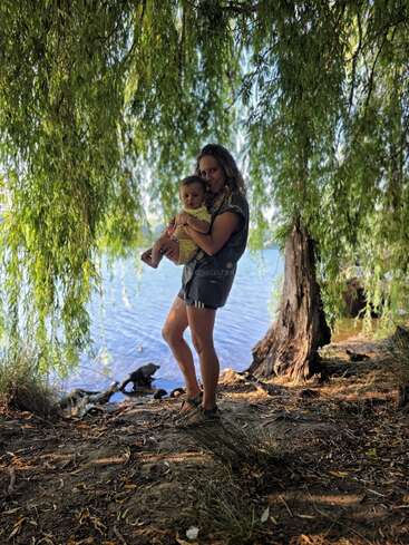 A woman holding a baby stands under a canopy of leafy green trees, near a serene lakeside, sunlight filtering through creating a peaceful, natural scene.