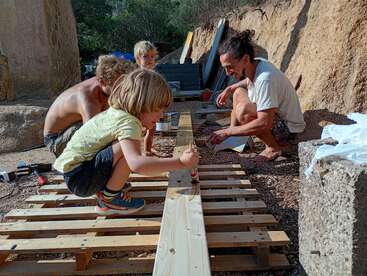 A group of children and adults are outdoors, working together on a DIY wood project, painting wooden planks, surrounded by nature, tools, and construction materials.