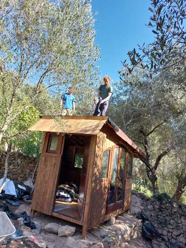 Two people stand on the roof of a small wooden cabin in a forest, surrounded by trees and rocks, working under a clear blue sky.