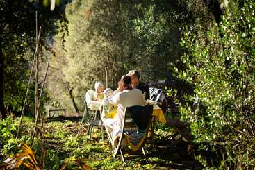 Una familia sentada en una mesa al aire libre, rodeada de árboles y vegetación, disfrutando de una comida soleada en un jardín tranquilo y natural, compartiendo risas.