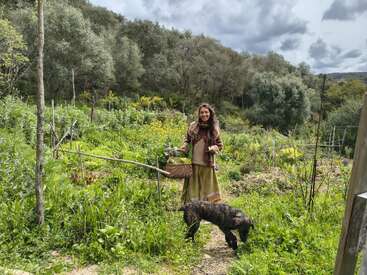 Uma mulher está em um jardim verdejante, segurando uma cesta de legumes, sorrindo, com um cachorro aos seus pés. Árvores e colinas cercam o cenário.