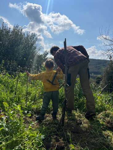 Uma criança e um adulto, talvez pai e filho, trabalham juntos ao ar livre num jardim exuberante e ensolarado, rodeados de vegetação sob um céu azul vibrante.
