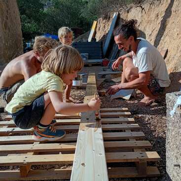 Um grupo de crianças e adultos está ao ar livre, trabalhando juntos em um projeto de madeira DIY, pintando tábuas de madeira, cercados pela natureza, ferramentas e materiais de construção.