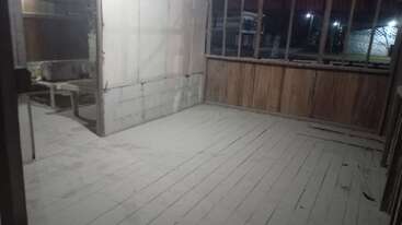 This image shows an empty, dusty wooden room with unfinished walls and open windows at night. A simple bench is visible in the corner near another room.