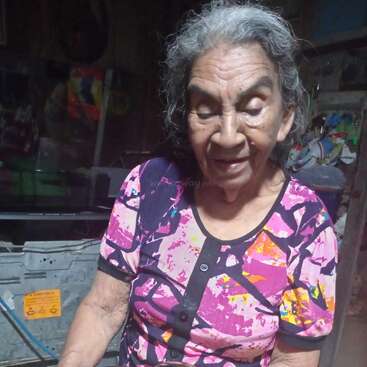 An elderly woman with gray hair and a colorful shirt sits at a wooden table, smiling, with a shiny metal pot placed in front of her.