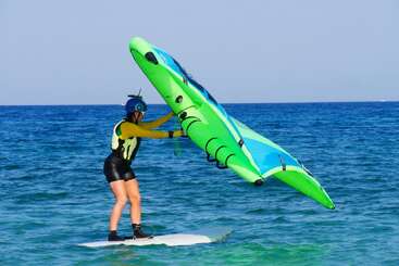 Una persona con casco y chaleco salvavidas hace equilibrios sobre una tabla en el océano, sujetando una vela hinchable de windsurf verde y azul en un día soleado.