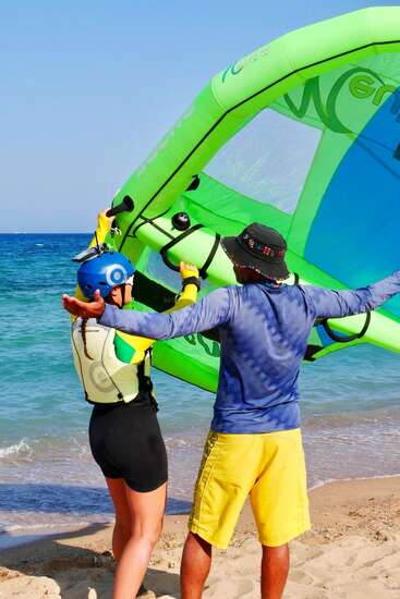 Dos personas de pie en una playa de arena, preparándose para el wing surfing. Una lleva casco y chaleco salvavidas, mientras la otra da instrucciones junto a un equipo verde.
