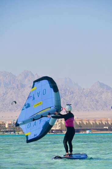 Una persona monta en una tabla hidroala, sujetando una gran ala azul en un día soleado y despejado. Al fondo se ven montañas, edificios y otros aficionados a los deportes acuáticos.