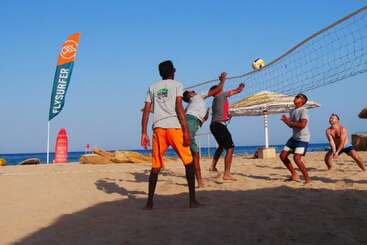 Un grupo de personas juega al voley playa bajo un cielo azul despejado. El océano, las sombrillas y una colorida bandera "FLYSURFER" se ven al fondo.