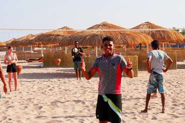 Un grupo de personas disfruta jugando al voleibol en una playa de arena, sonriendo bajo la brillante luz del sol, con sombrillas de paja y el cielo azul de fondo. Predominan las vibraciones veraniegas.