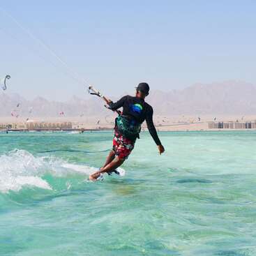 Un hombre practica kitesurf en aguas turquesas, con traje de neopreno y gorra. Varios otros kitesurfistas y cometas de colores son visibles con las montañas del desierto en el fondo.