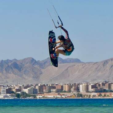 A person is kiteboarding high above the water, performing a trick. Mountains and a city skyline are visible in the background under a clear blue sky.