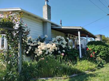 Une charmante maison blanche avec une cheminée, entourée de fleurs et de verdure luxuriante sous un ciel bleu clair, dégageant une atmosphère paisible et rurale.
